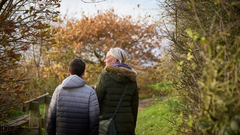 Two people walking down a narrow path framed by dense shrubs and trees, with autumn leaves visible ahead and water in the distance.